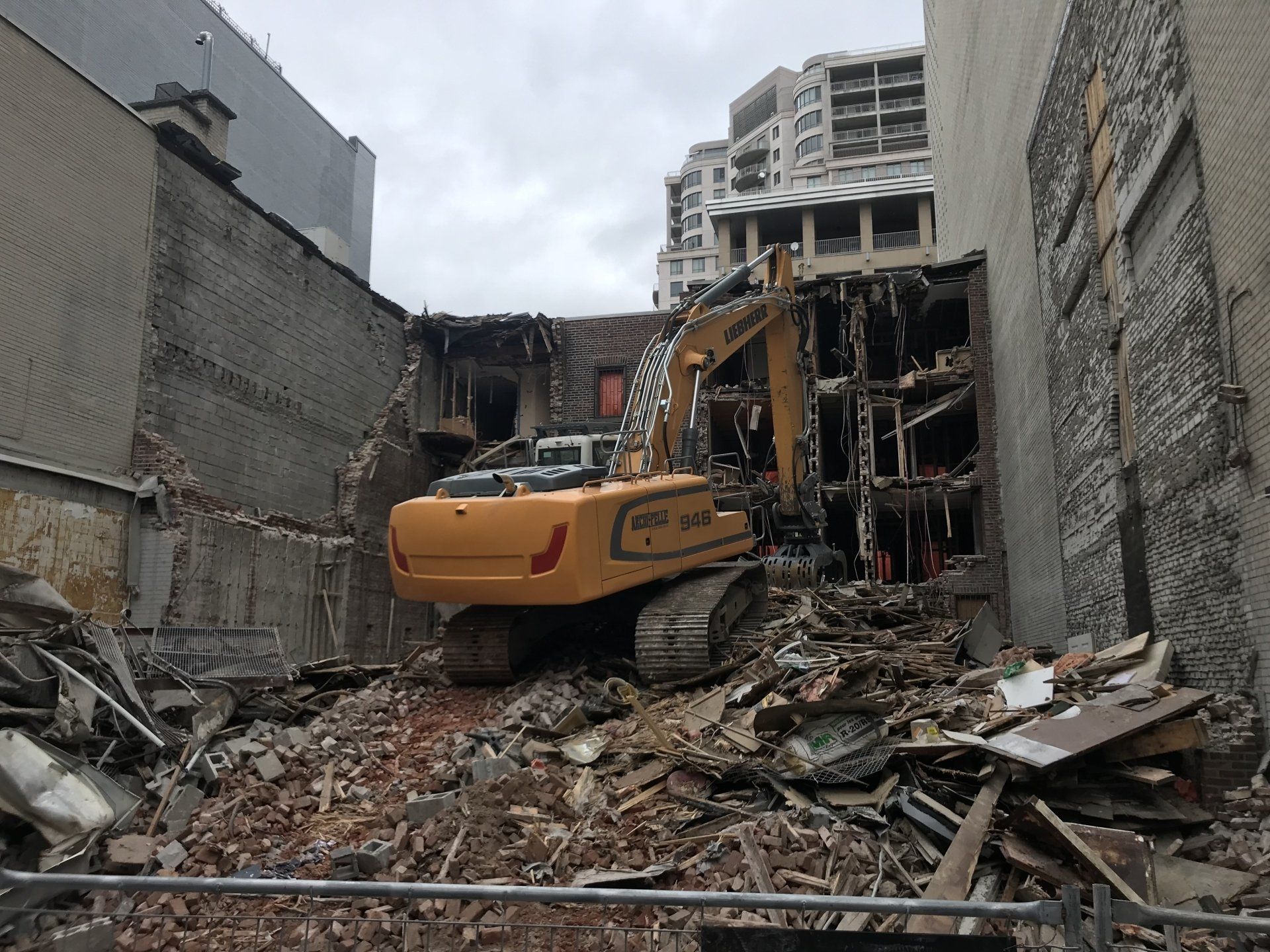 Chantier de démolition avec une excavatrice entourée de gravats, de murs de briques et d'un bâtiment à plusieurs étages.