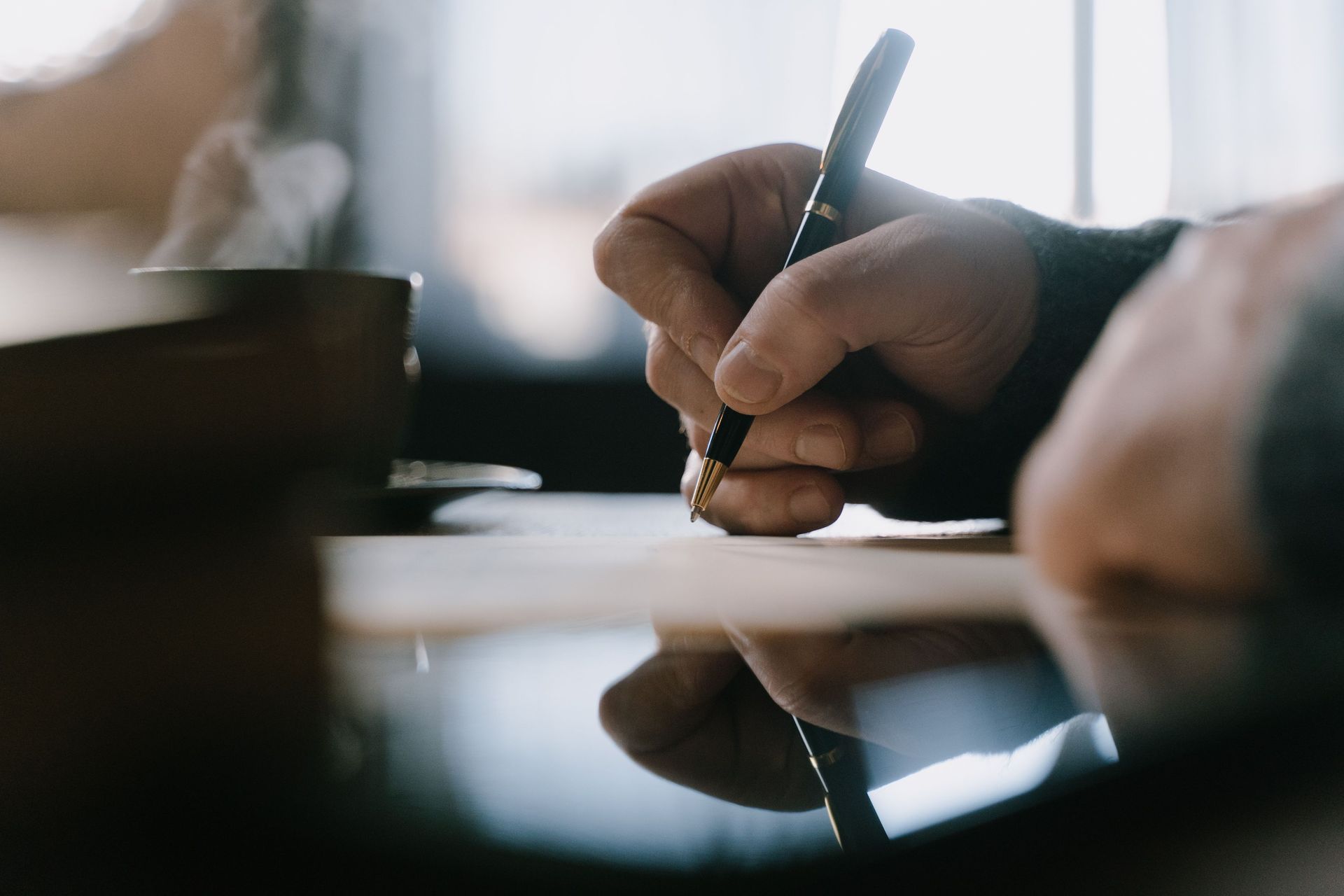 Hand writing with a pen at a table, with a cup blurred in the background