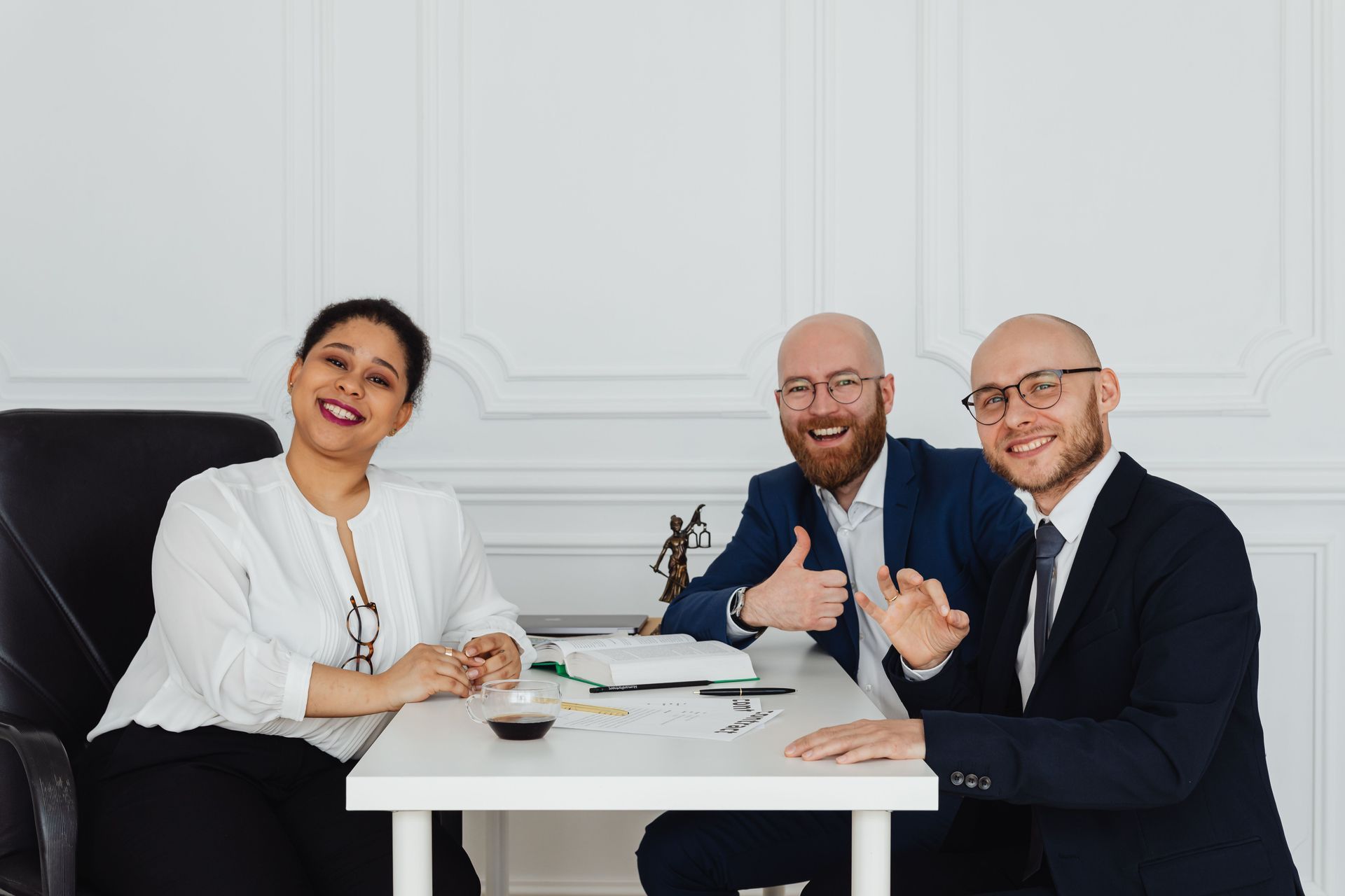 Three professionals smiling at a white table in a bright office, discussing documents and a laptop.