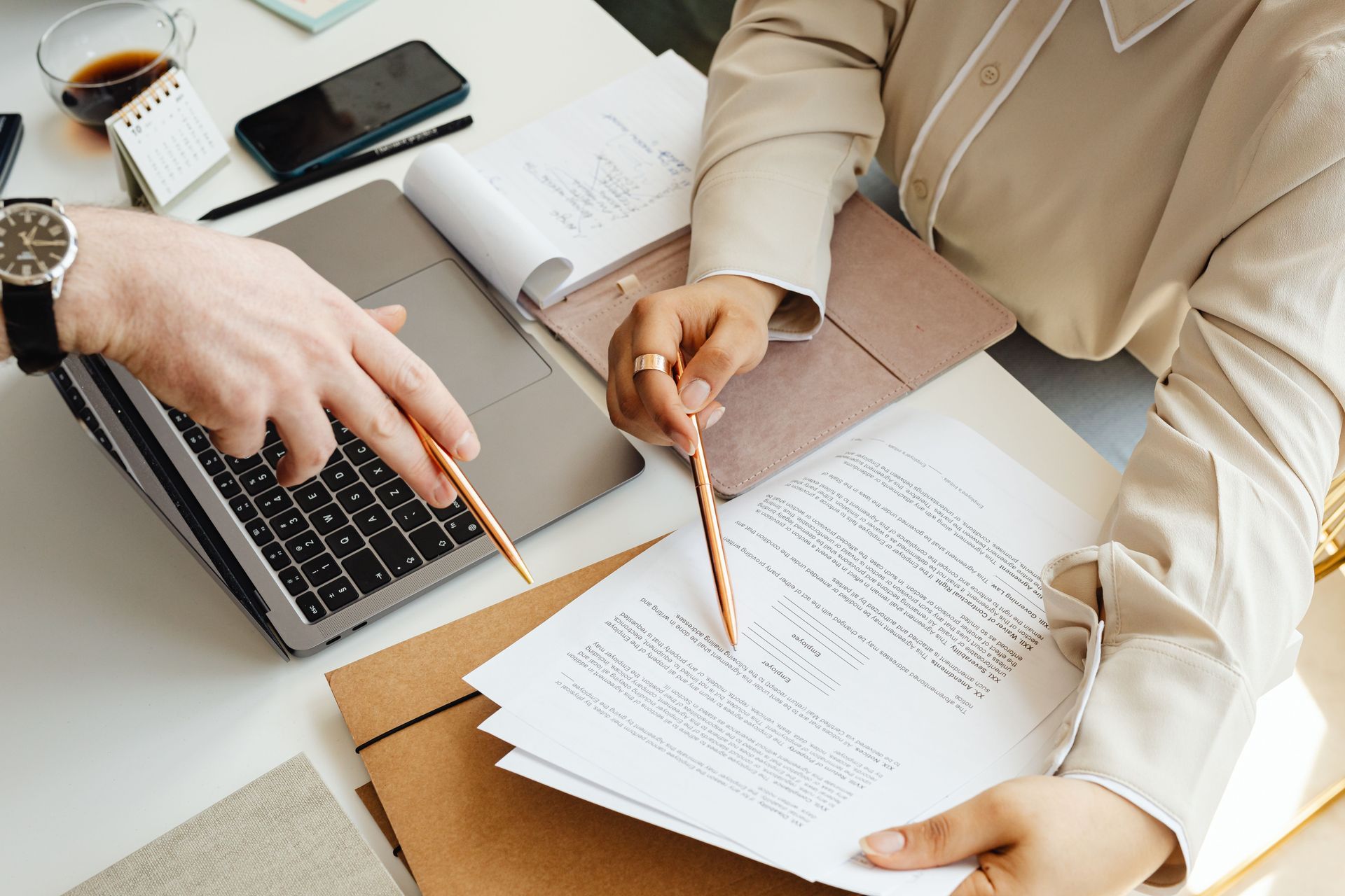 Two people reviewing documents at a desk with a laptop, coffee, and pencil holders