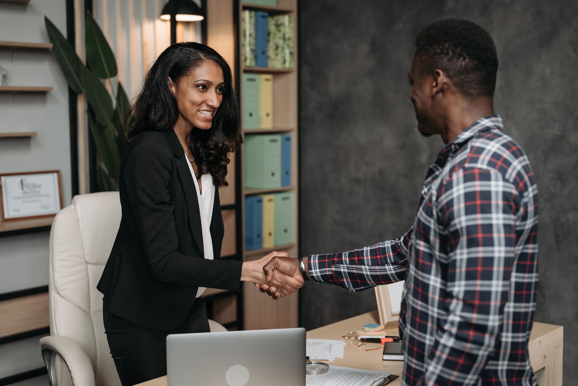 Two colleagues shaking hands across an office desk, with a laptop and shelves in the background.