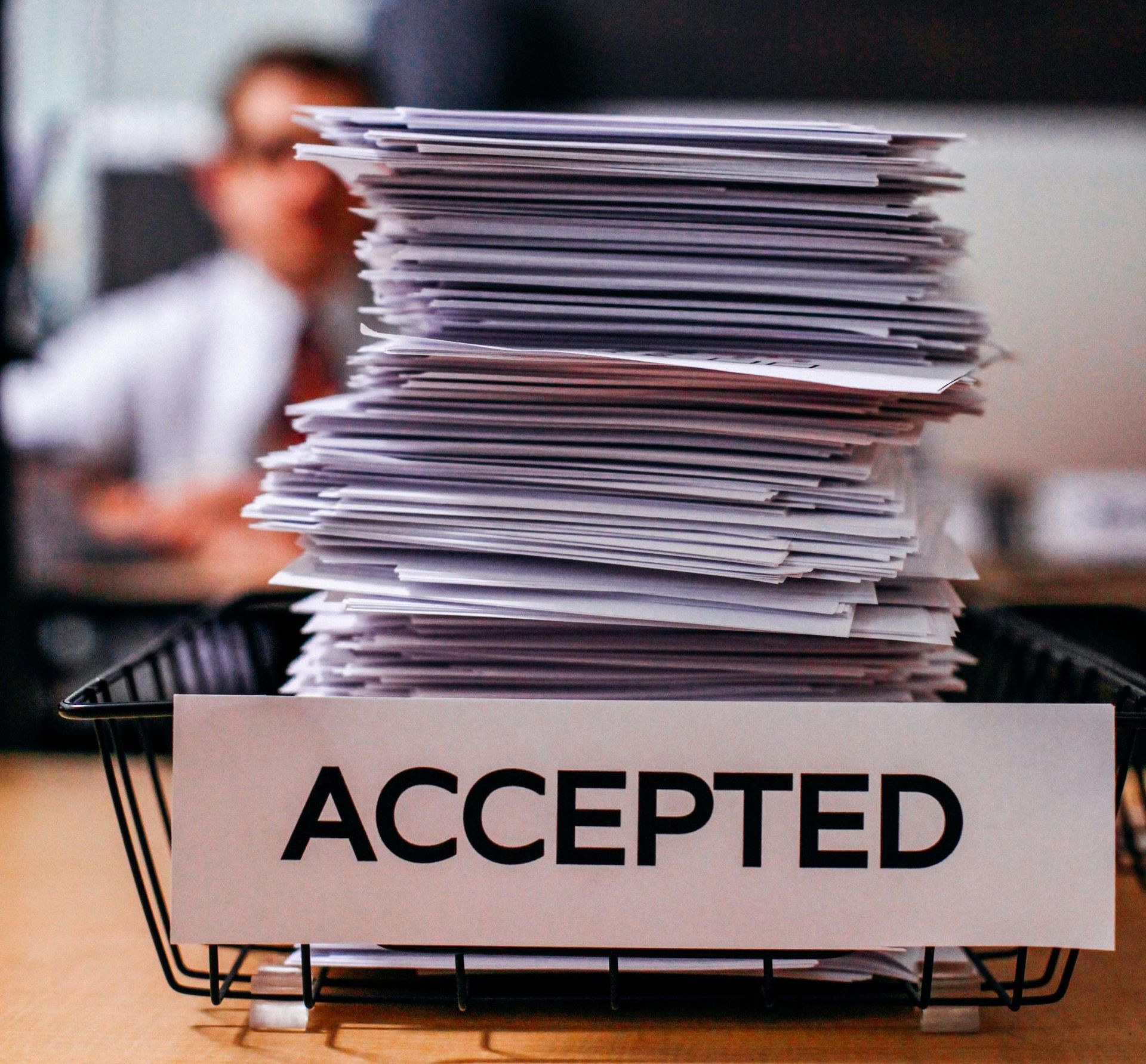 Stack of paperwork in a wire tray labeled “ACCEPTED” on a desk, with a blurred office worker in the background