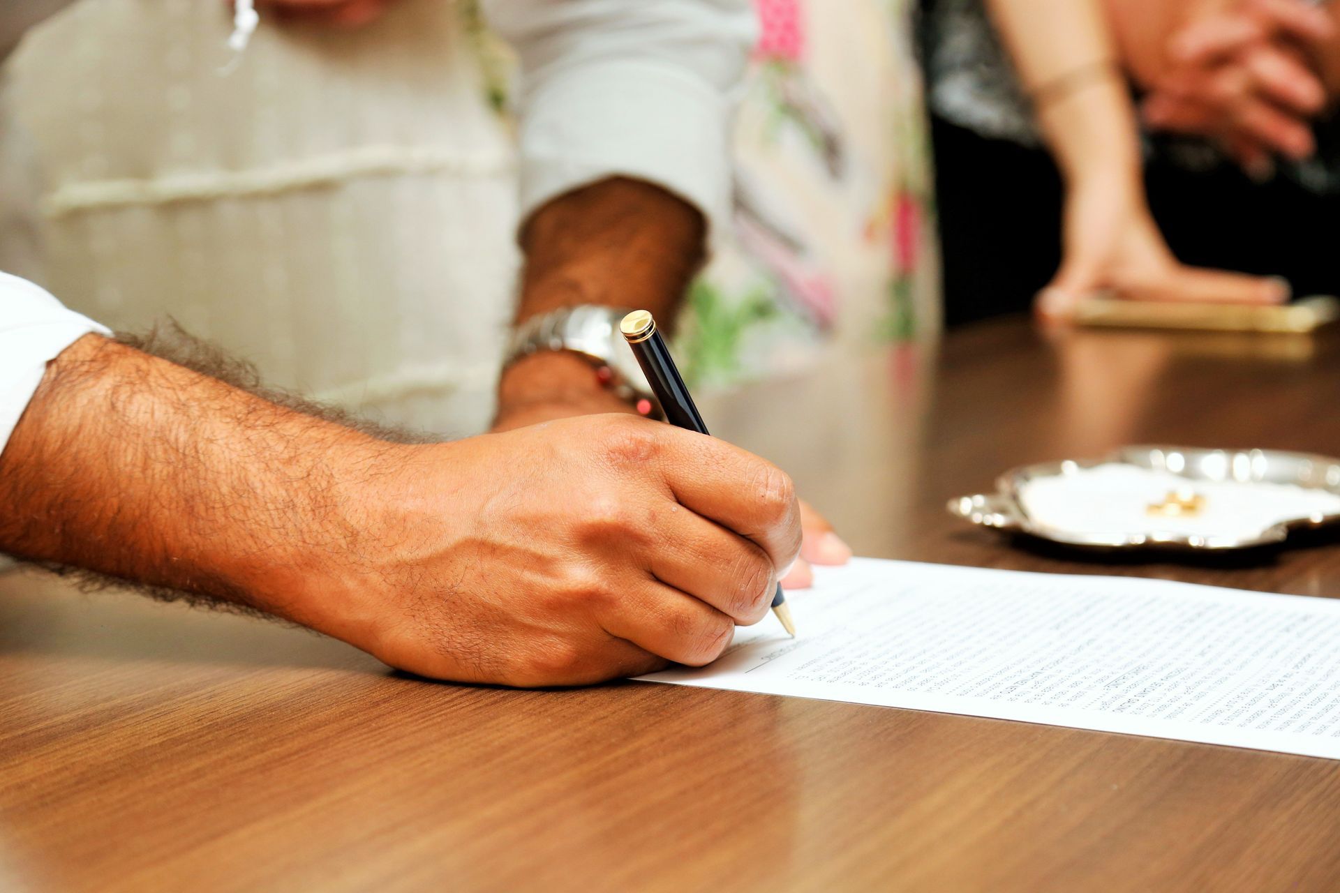 Hand signing a document on a wooden table, with a pen and plate in the background