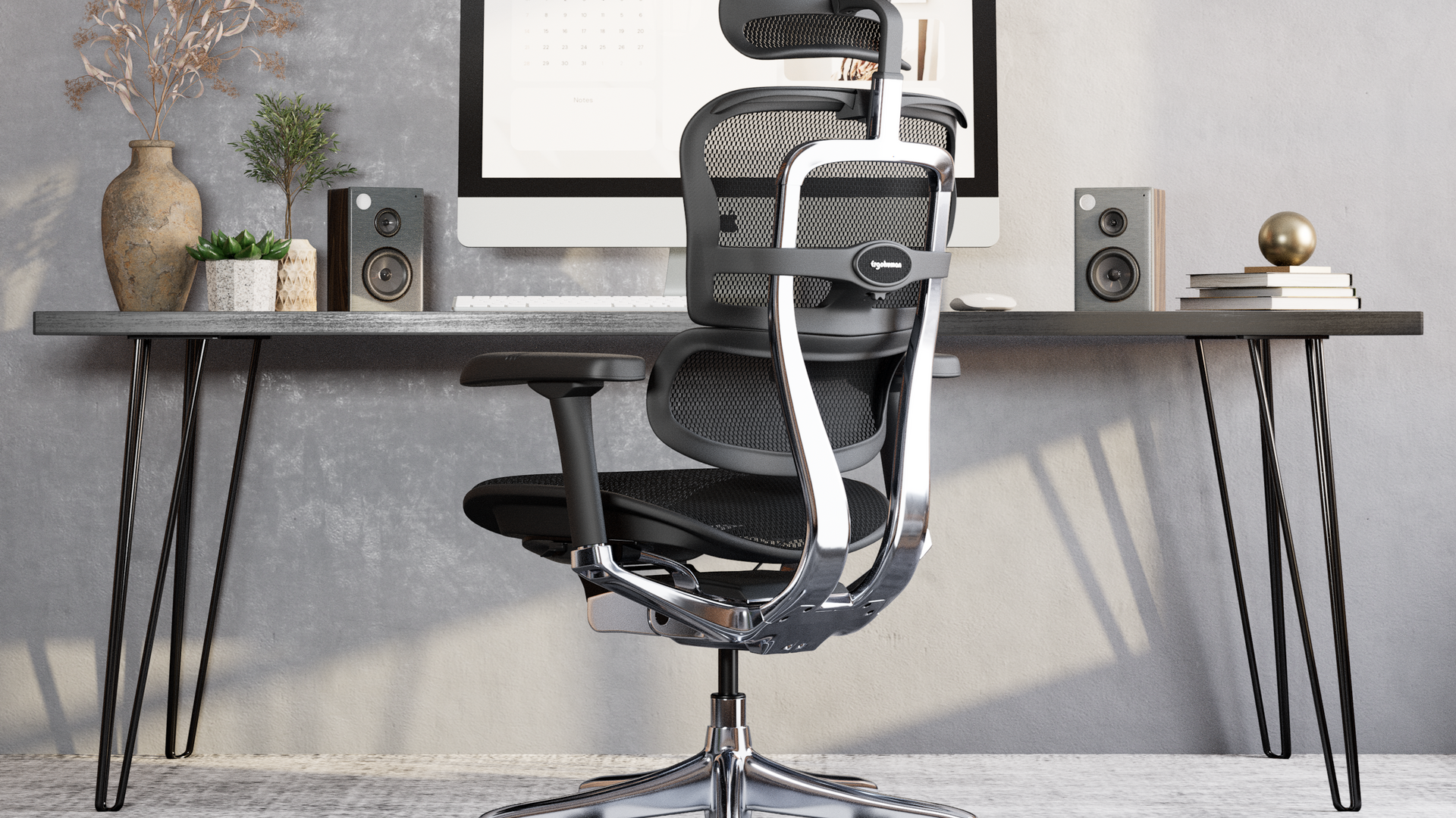 Black Ergohuman Elite office chair situated at a black desk with hairpin legs. The carpet and walls are grey. On the desk is an iMac, speakers, plants and books