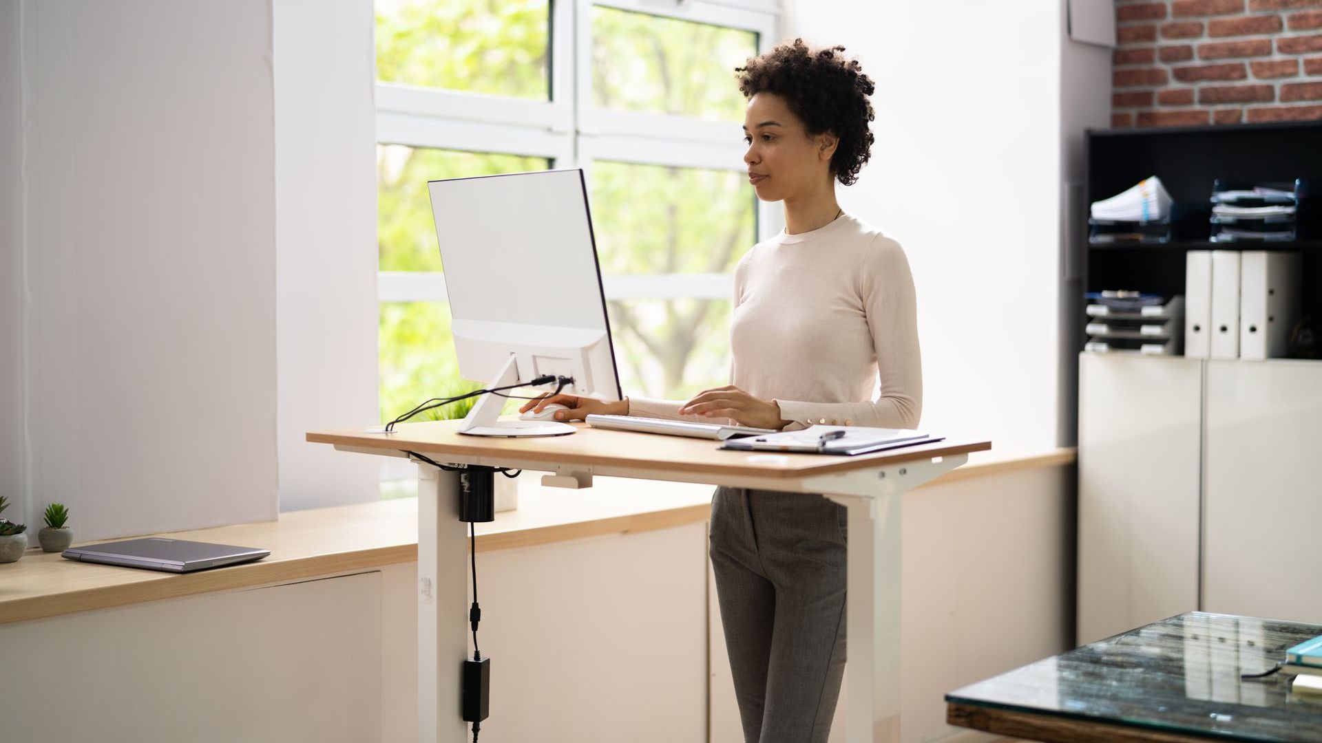 A woman wearing a white long-sleeved top and grey trousers is standing at a height-adjustable desk. On the desk is a monitor, keyboard and a pile of papers with a pen on top. In the background is an office with natural daylight, a table, and filing cabinets.
