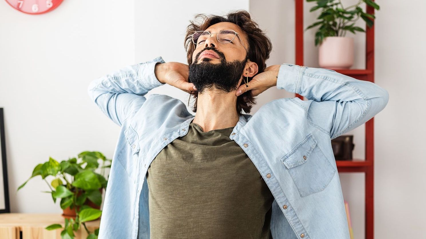 A man wearing a green t shirt and denim overshirt, leaning back in his chair, stretching his back and arms behind his neck. He has a beard and glasses.