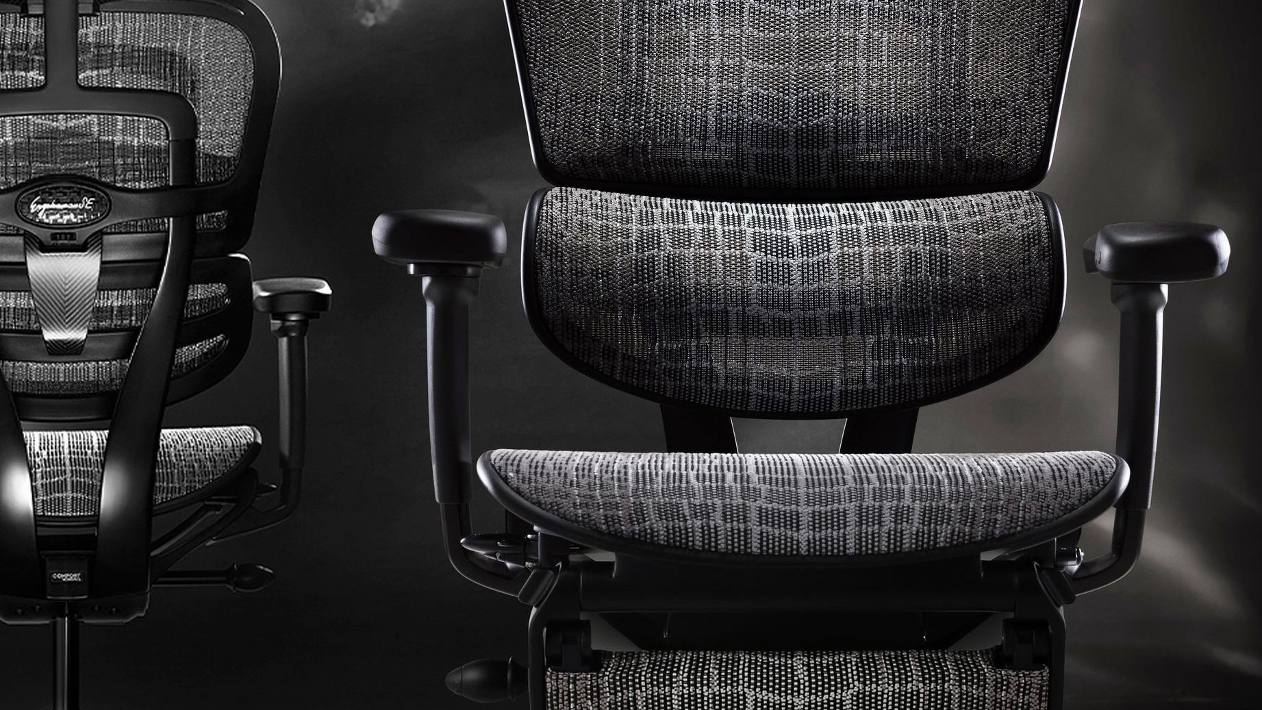 Two Ergohuman Carbon gaming chairs against a black smokey background. One chair is facing the camera, and the other is facing away.