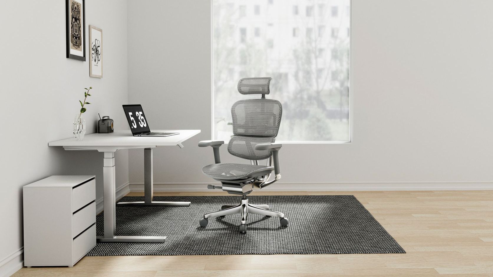 Grey Ergohuman chair on a black rug at a white desk looking at a laptop. There is a white wall and a soft window behind the chair.