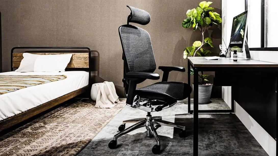 Black mesh office chair facing at a desk in a bedroom. On the desk there is a monitor and a small amber bottle. In the background is a healthy green plant.