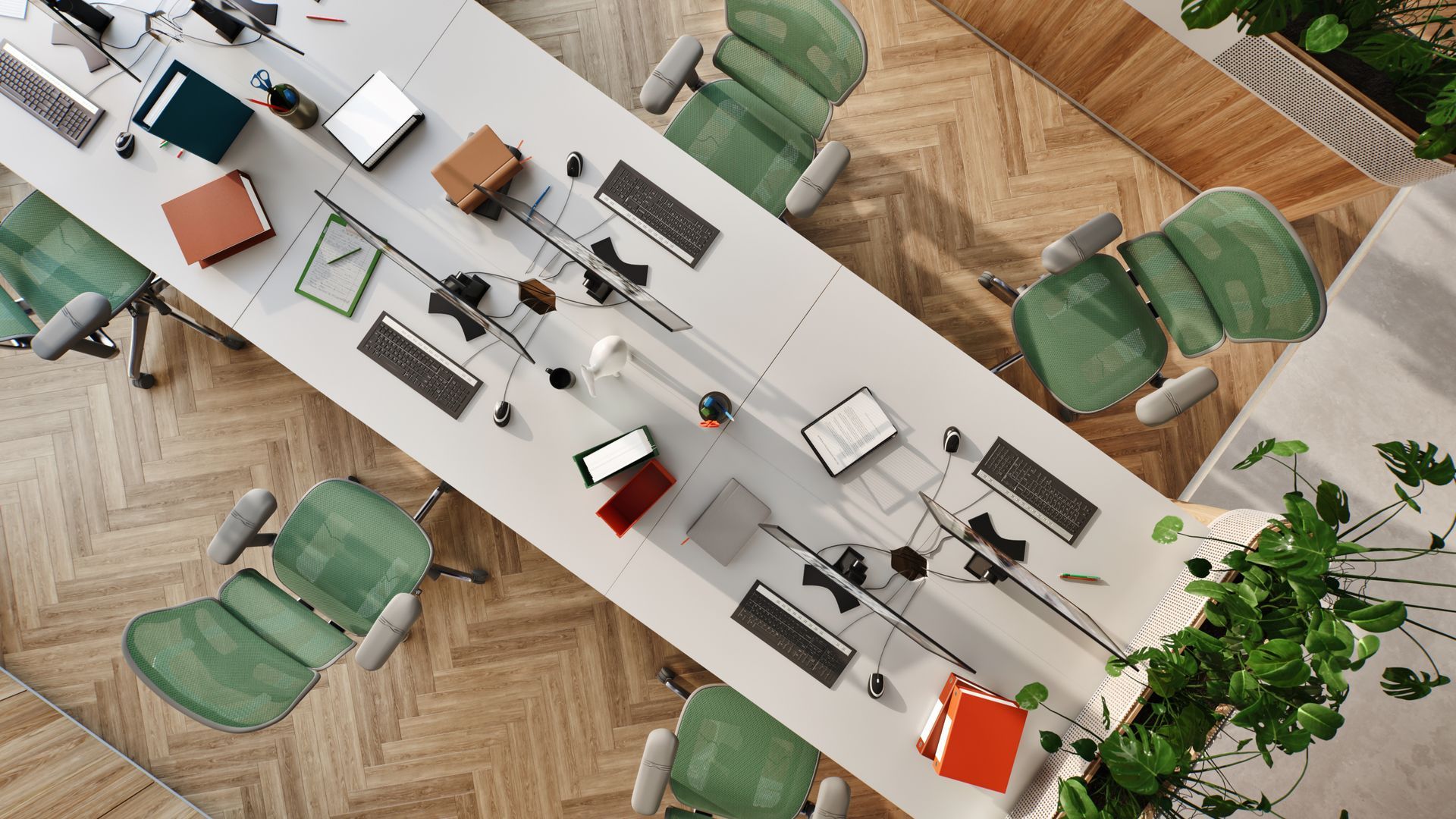 Birdseye view of an office with a white desk in the middle, with six chairs at each section of the large desk. The chairs are green mesh Ergohuman office chairs in a grey frame. At each desk is a computer, keyboard, notebooks, and some books. The floor is wooden herringbone. The office is adorned with green plants. 