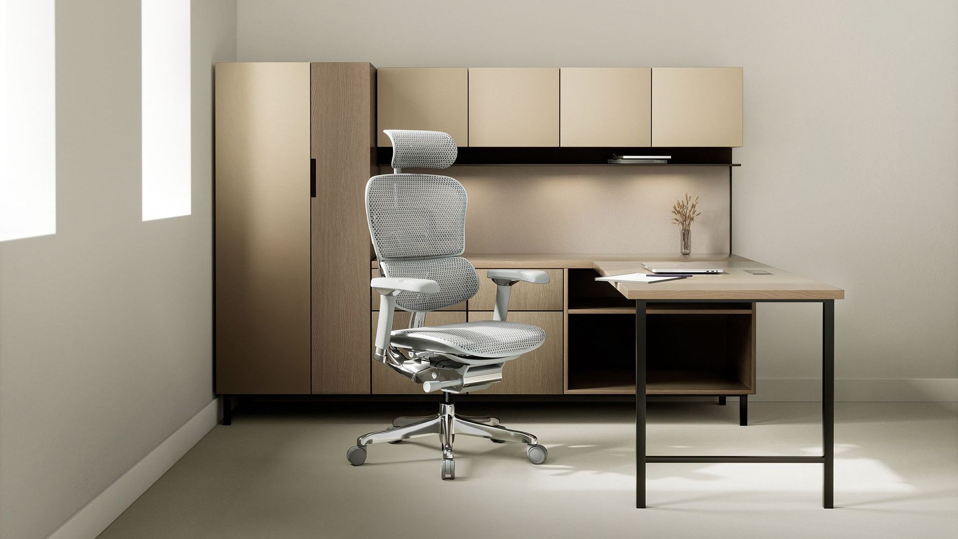 Grey Ergohuman chair on a black rug at a white desk looking at a laptop. There is a white wall and a soft window behind the chair.