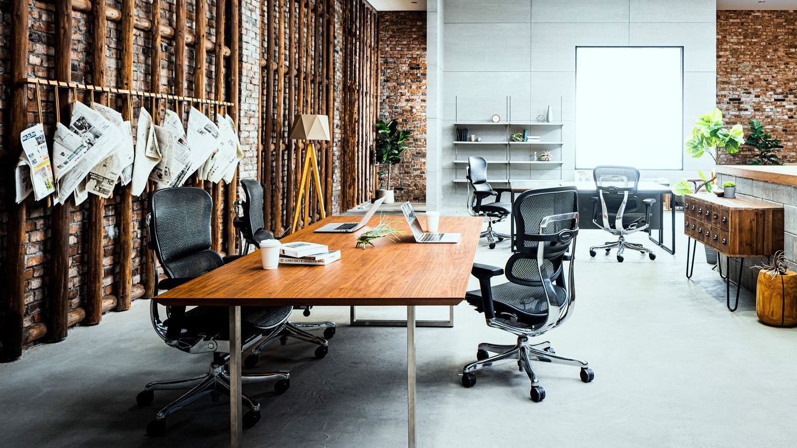 Three black Ergohuman office chairs at a wooden table. There are laptops, books and takeaway cups on the table. There is another table with Ergohuman chairs in the background.