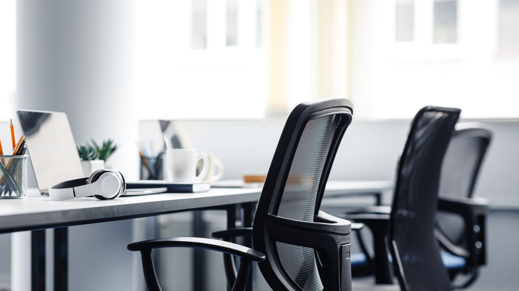 A few mesh office chairs facing a grey desk. On the desk are laptops, mugs and headphones.