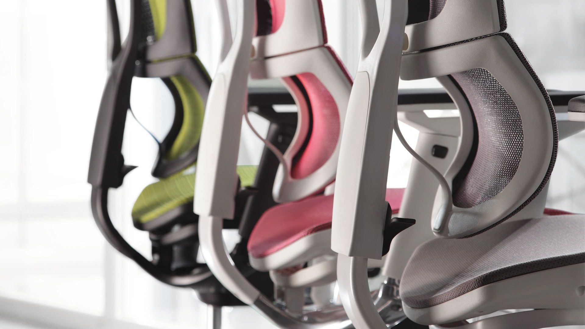 3 Ergohuman chairs facing a desk. We see the back of the chairs. There is a grey chair, pink and green.