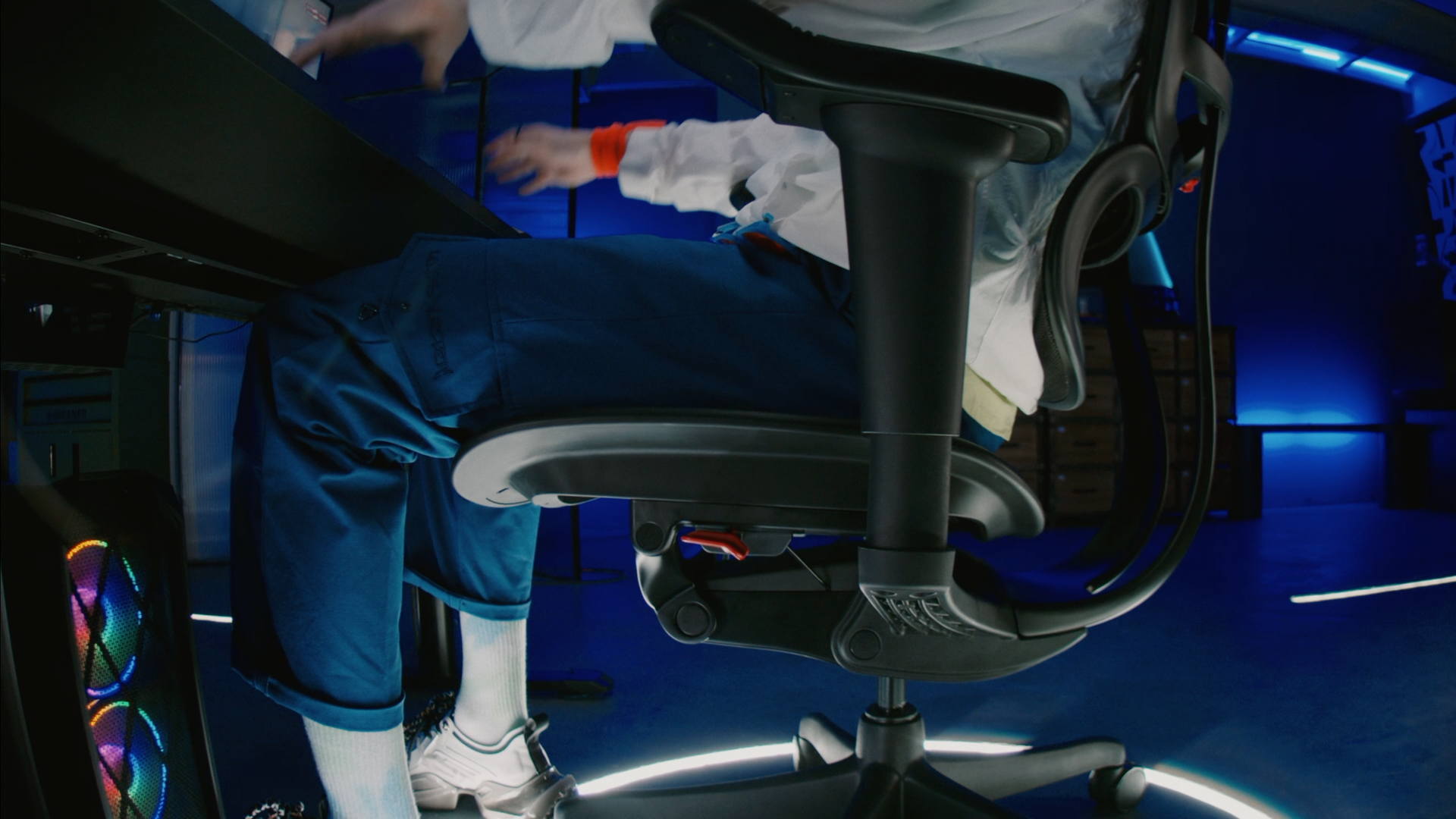 Black Ergohuman gaming chair (Ultra) profile view with a person sitting in the seat. The person is wearing blue trousers, white socks and a white jacket. They are playing a game at the desk.