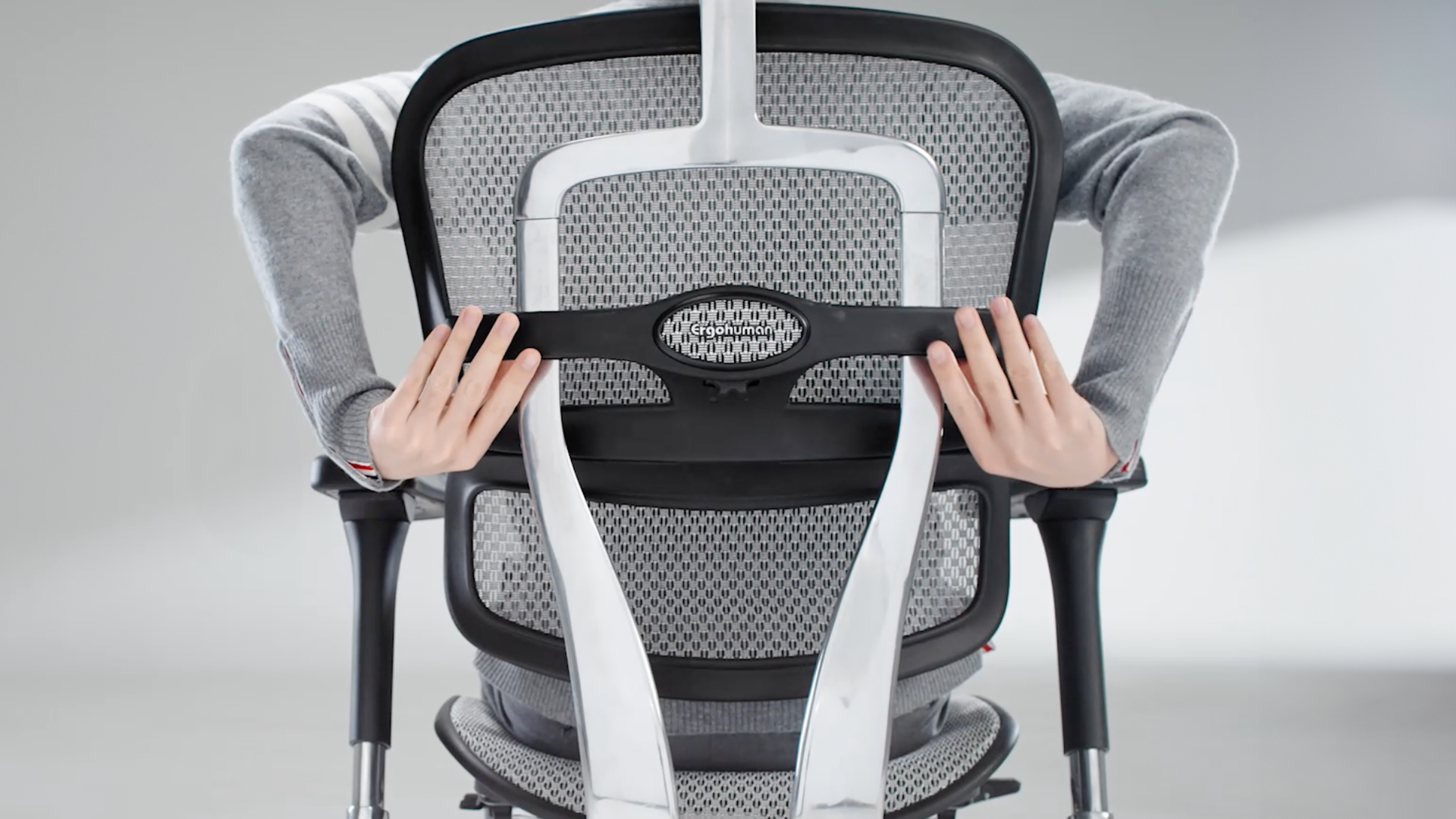 The back of the Ergohuman office chair in a black frame with grey mesh. The man sitting in the chair has his arms behind the chair to show how easy it is to adjust the back while sitting.