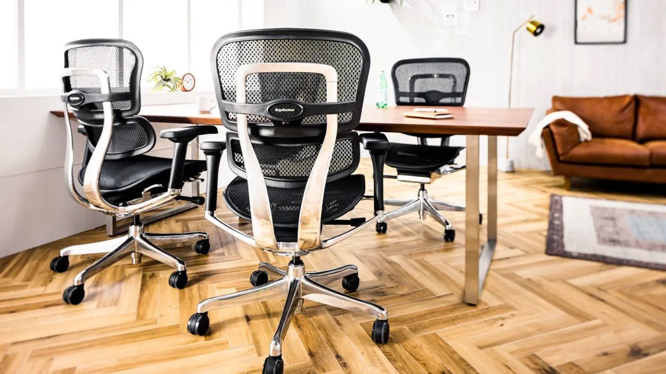 Four Ergohuman Elite chairs in black frame with black mesh around a wooden table. The floor is herringbone wood. There is a brown sofa in the background.