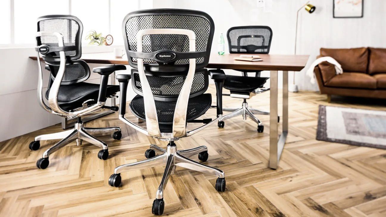 Four Ergohuman chair's at a wooden desk. There is a brown sofa in the background behind a rug. Natural light is streaming in from the window at the left.