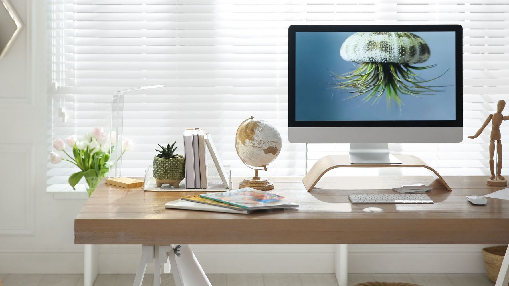 Wooden deks at a window. On the desk is a monitor stand to reduce eye strain and a desktop. On the desk is also a wooden figure, keyword, magic mouse, globe, and a collection of books.