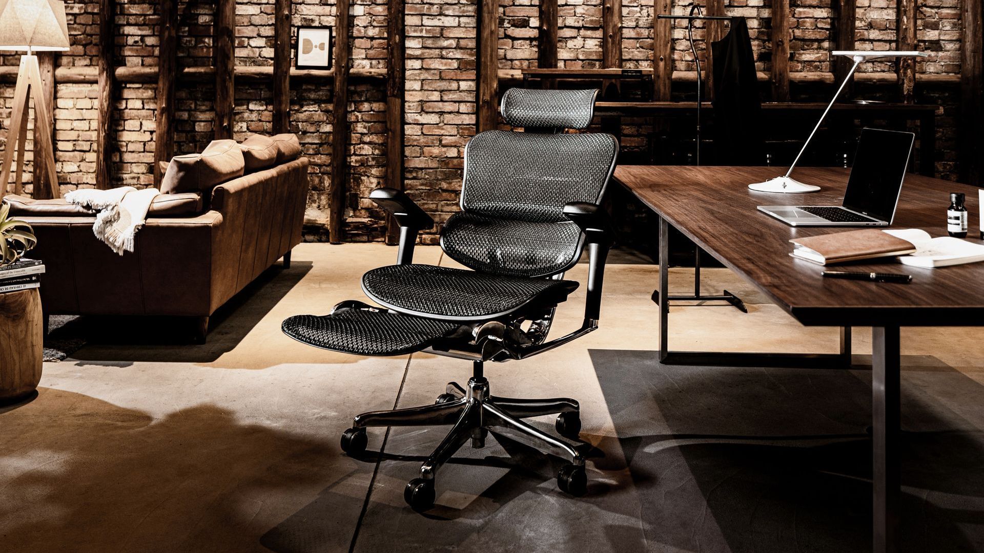 Black mesh ergonomic office chair in a relaxed lounge space with a desk and leather sofa.
