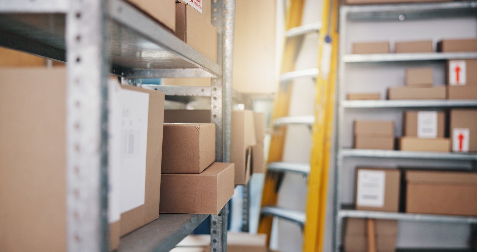 Storage room shelves stocked with cardboard boxes beside a yellow ladder.
