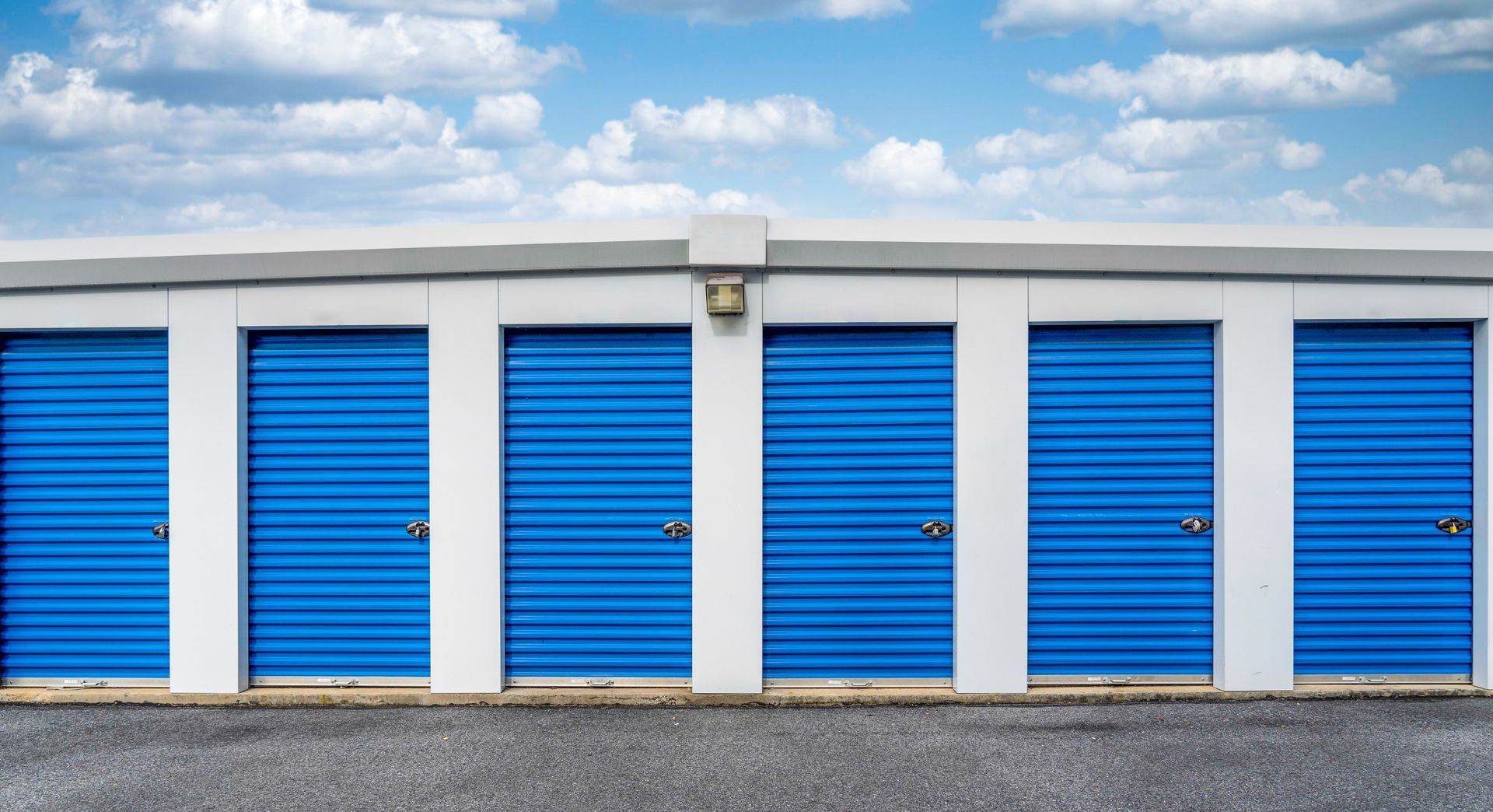 Row of blue roll-up doors on outdoor self-storage units under cloudy sky.