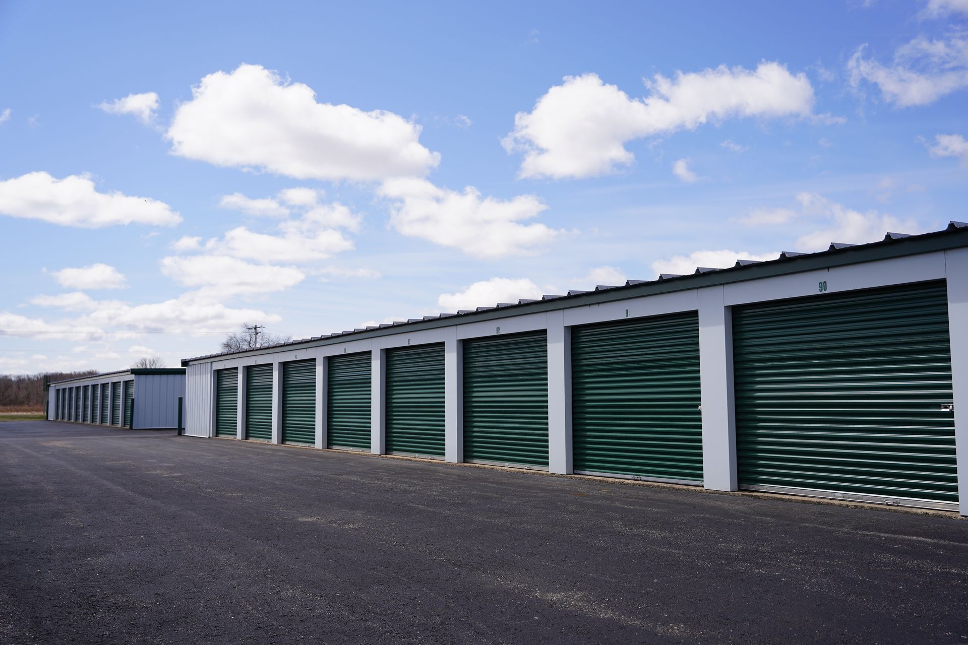 Row of outdoor storage units with numbered green roll-up doors under a cloudy sky.
