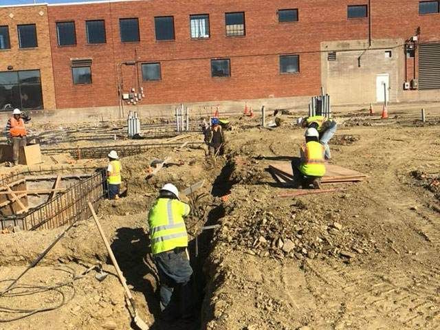 A group of construction workers are digging in the dirt in front of a brick building. A group of construction workers are digging in the dirt in front of a brick building.