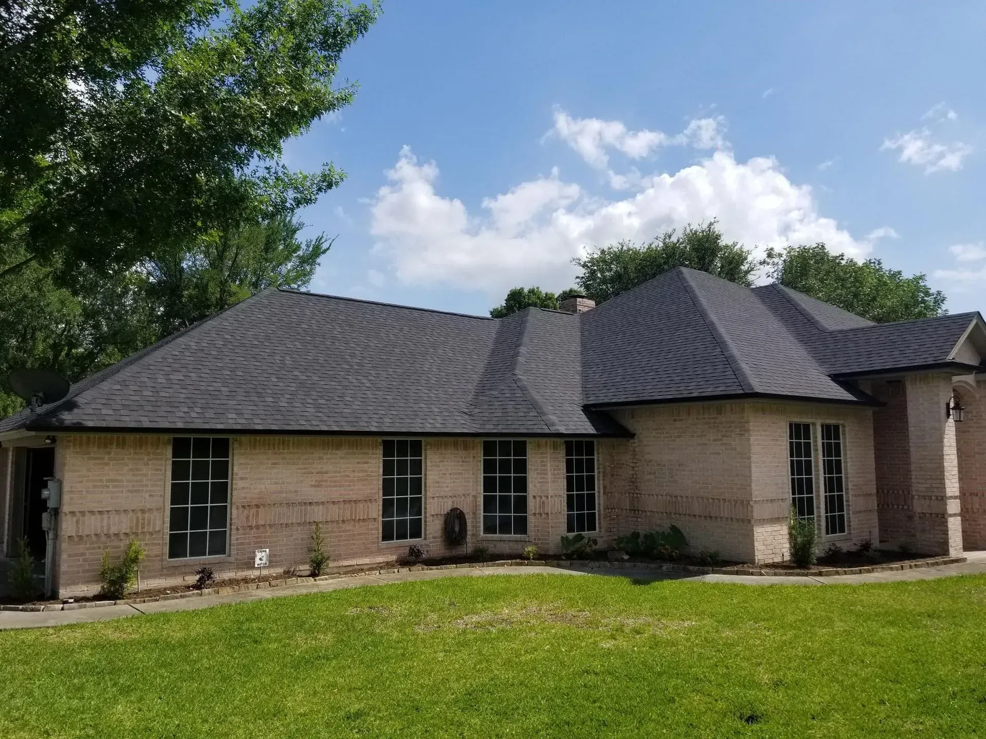 Tan brick house with dark gray shingle roof, green lawn, and blue sky with clouds.