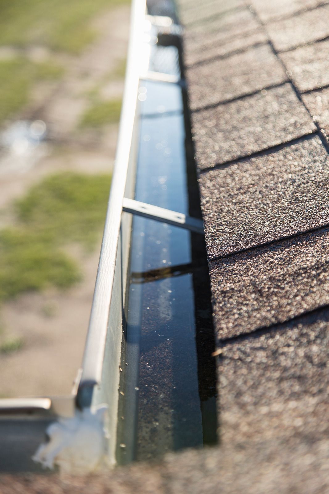 Close-up of a rooftop gutter beside brown shingles, with a blurred yard in the background