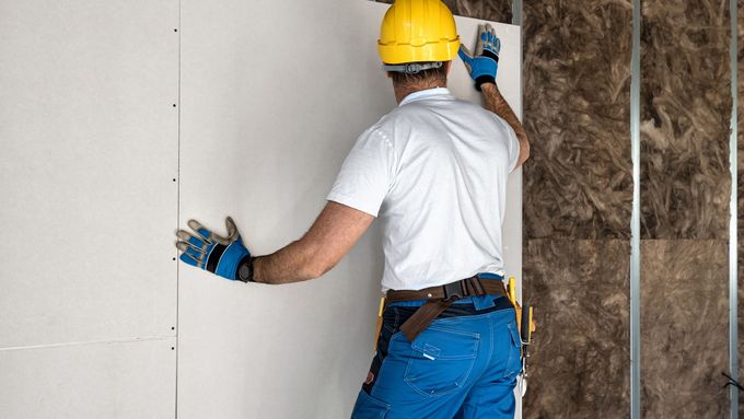 Worker in yellow hard hat installing wall insulation panel with blue gloves.