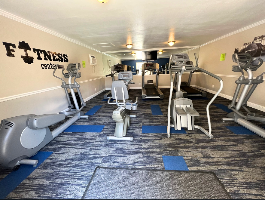 Gym with various exercise machines: elliptical, recumbent bike, treadmill. Blue and gray carpet.