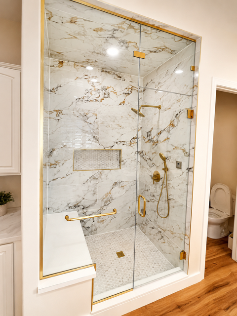 Walk-in shower with marble-look tile, glass doors, brass fixtures, and a built-in bench