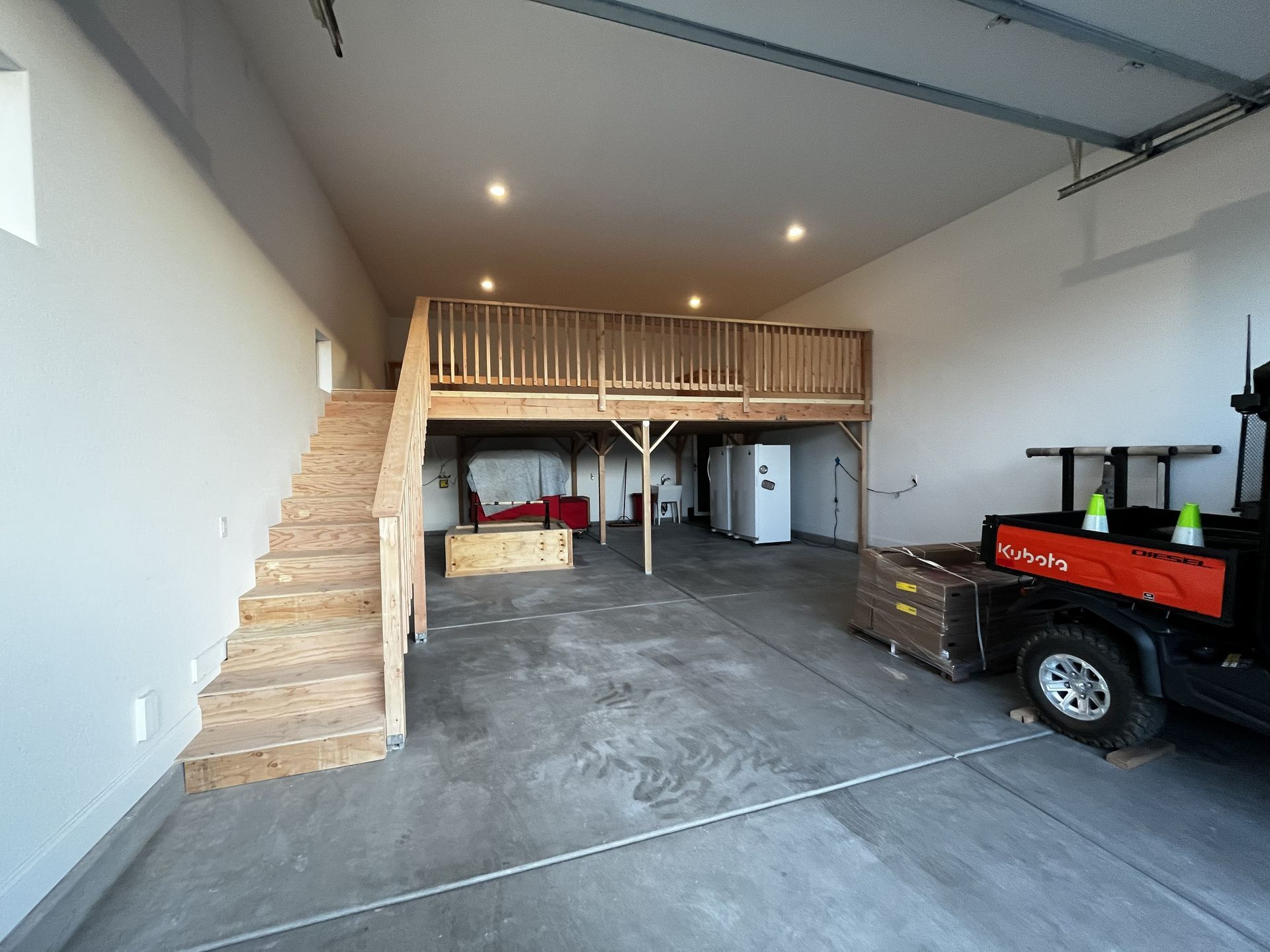 Spacious garage with loft storage, wooden stairs, concrete floor, and a red utility trailer.