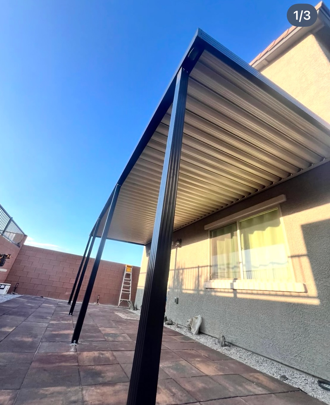 Covered patio with corrugated metal awning beside a beige house and brick-paved courtyard