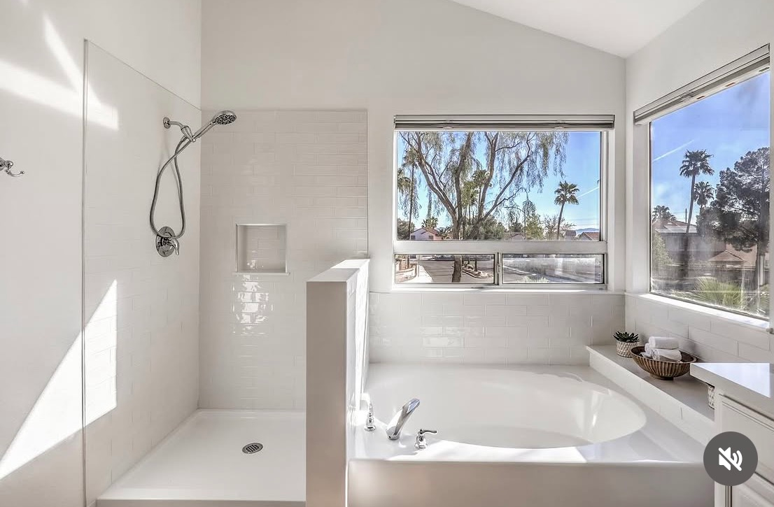Bright white bathroom with shower, soaking tub, and large windows overlooking trees.