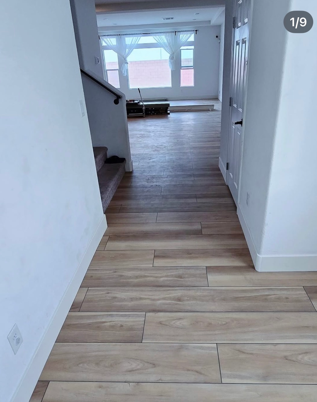 Narrow hallway with light wood floors leading to a bright living room and staircase on the left.