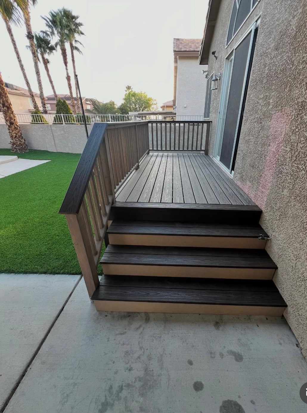 Short outdoor stairway to a raised wooden deck beside a house with a black railing.