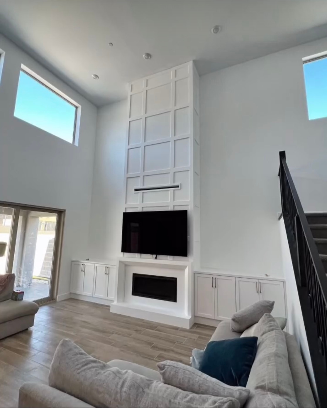 Bright living room with tall white fireplace, high windows, gray couch, and staircase
