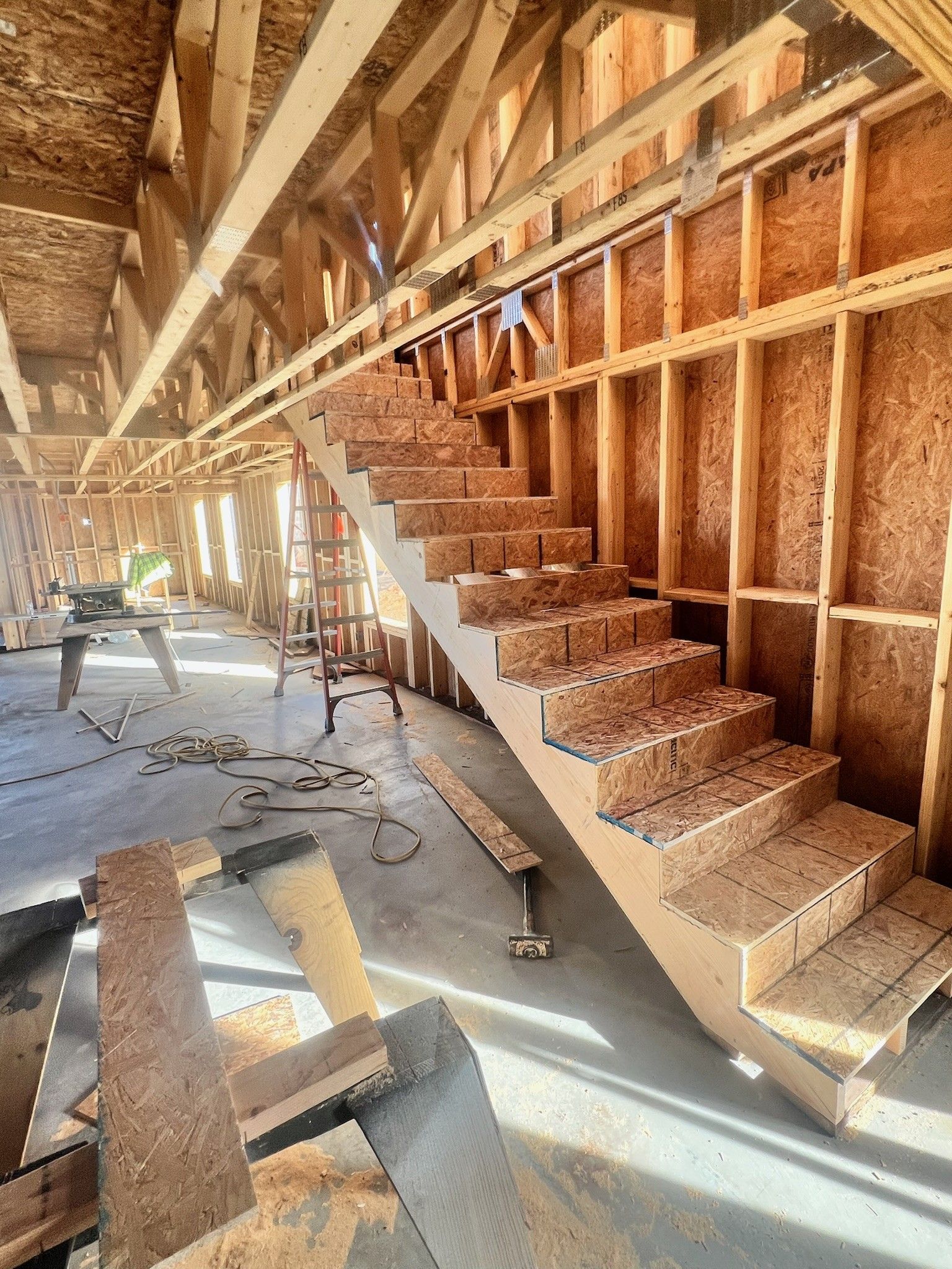 Unfinished wooden staircase inside a framed building under construction, with exposed studs and sunlit floor.