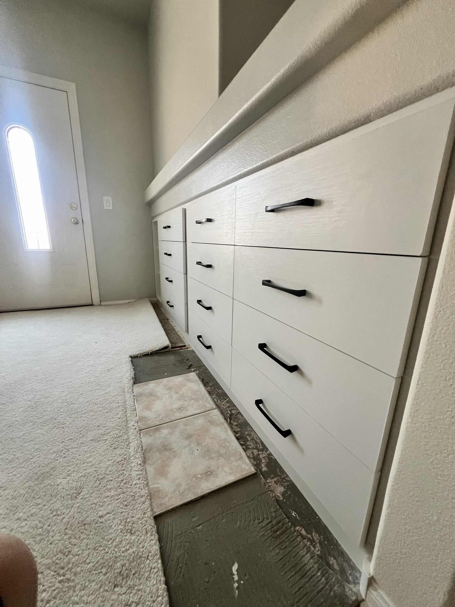 White built-in drawers and cabinets along a carpeted hallway beside a bright door.