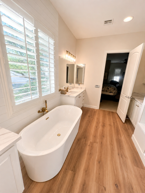 Bright modern bathroom with freestanding tub, vanity, large window shutters, and wood-look floor.