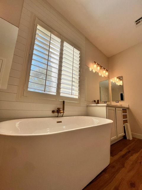 Bright modern bathroom with freestanding tub, window shutters, vanity, and warm wall sconces.