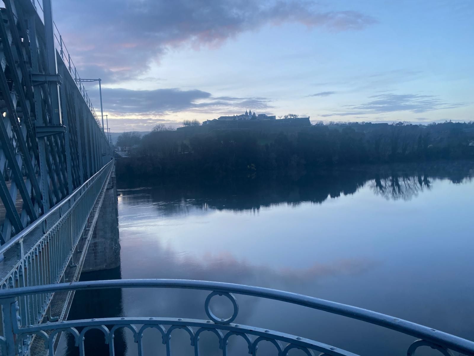 Vista de un lago nevado al atardecer desde un puente, con el agua en calma reflejando el cielo azul y los árboles.