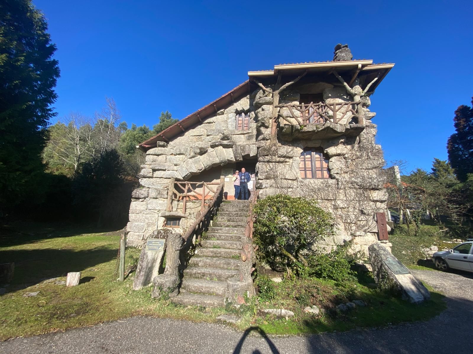 Casita de piedra con escaleras empinadas y un pequeño balcón, situada en un soleado patio con césped y árboles.