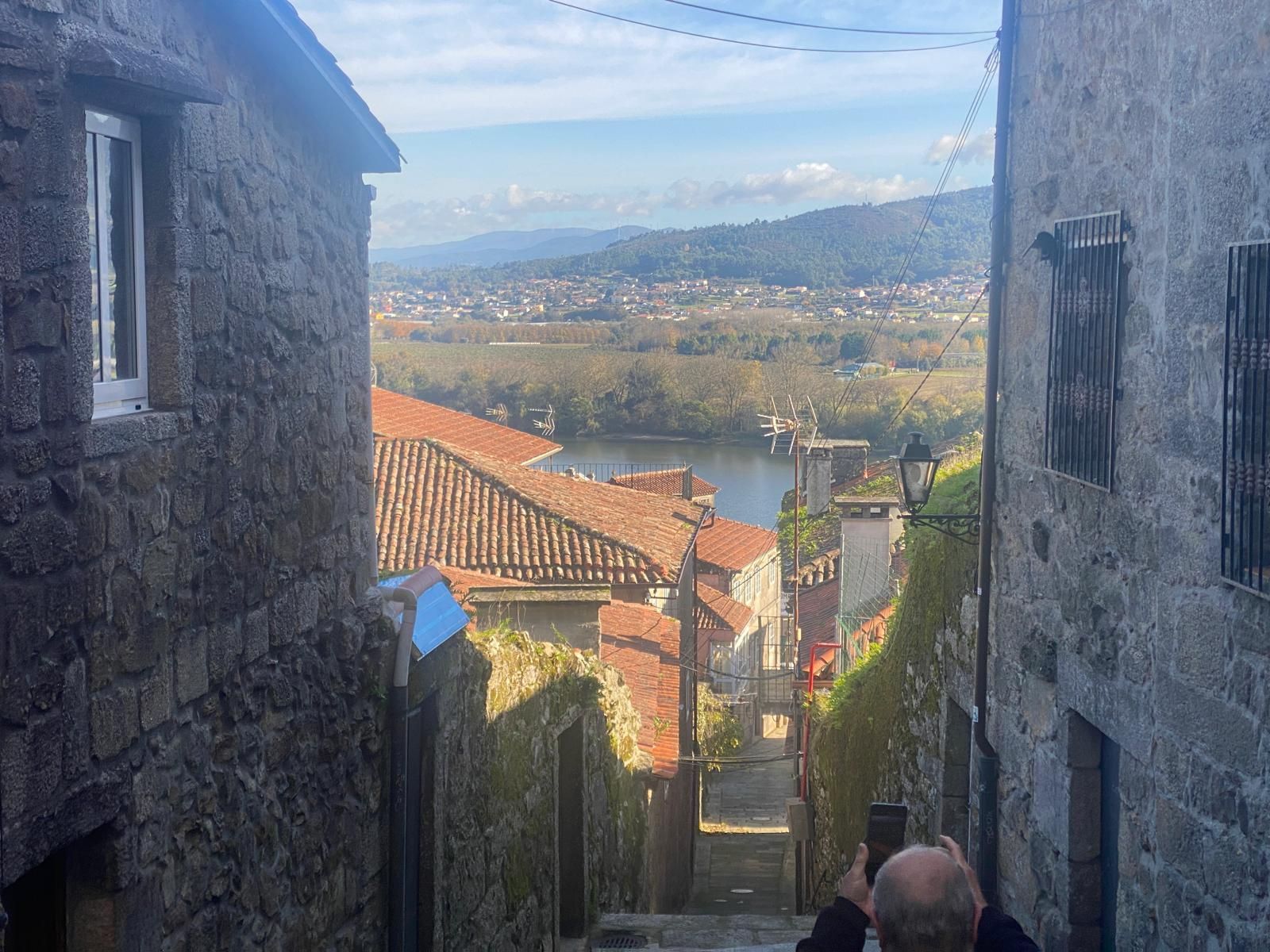 Estrecho callejón de piedra que desciende hacia un pueblo de tejados rojos con valle y montañas al fondo.
