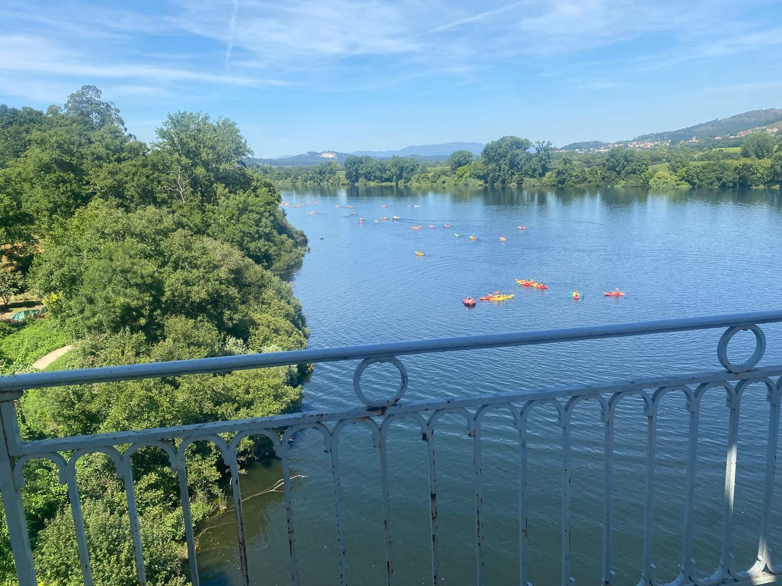 Lago azul con kayaks, visto desde un puente sobre árboles y colinas bajo un cielo despejado.