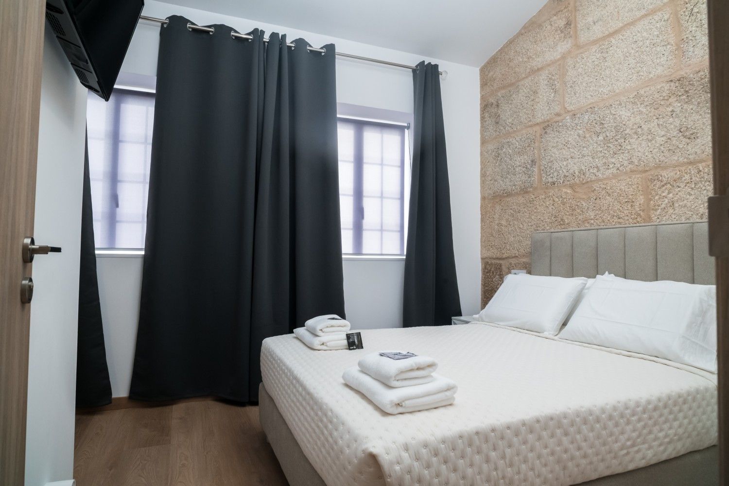 Habitación luminosa con cama, toallas dobladas, cortinas oscuras y una pared decorativa de piedra.