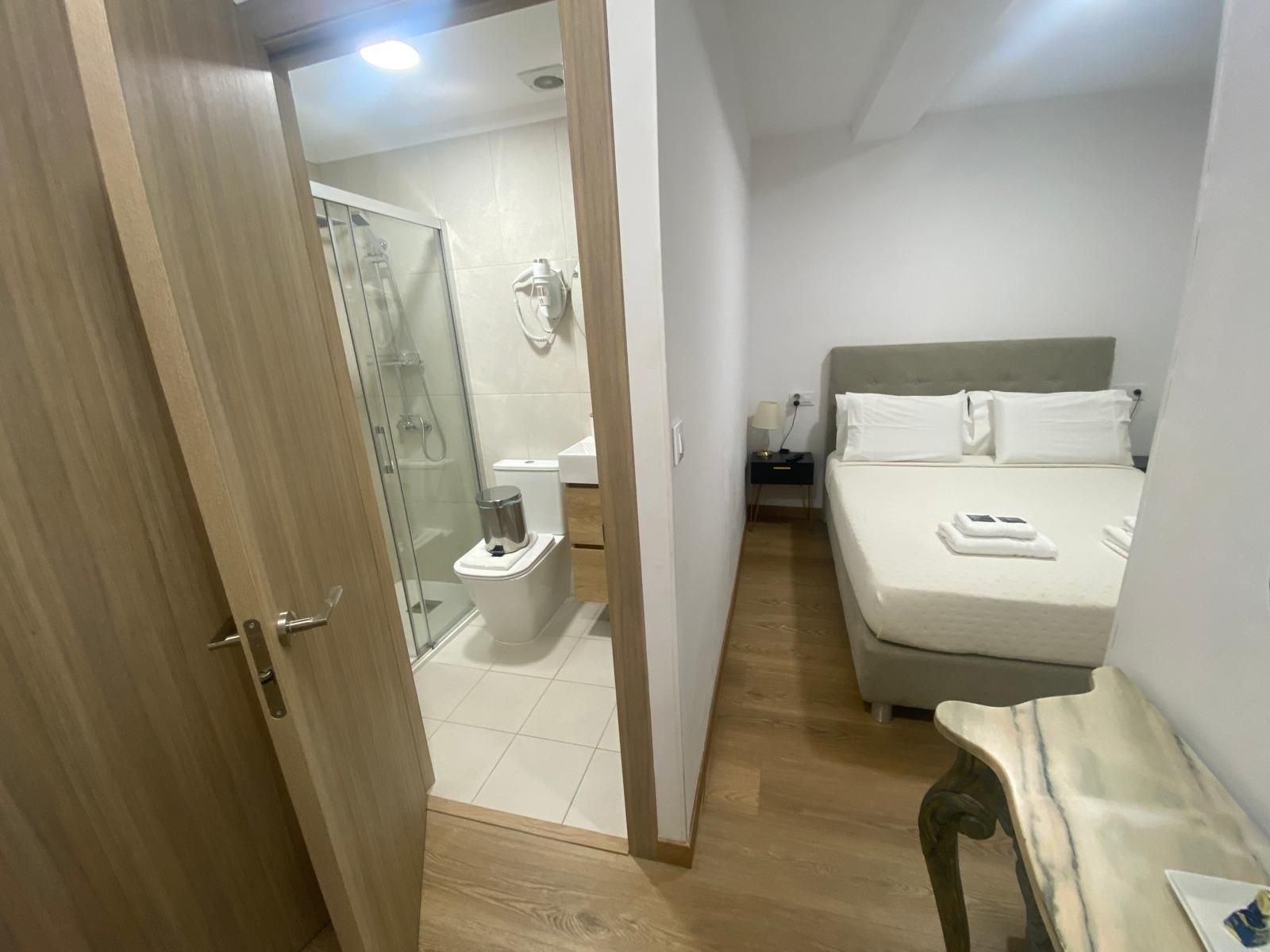 Habitación de hotel con la puerta del baño abierta, cama blanca y suelo de madera.