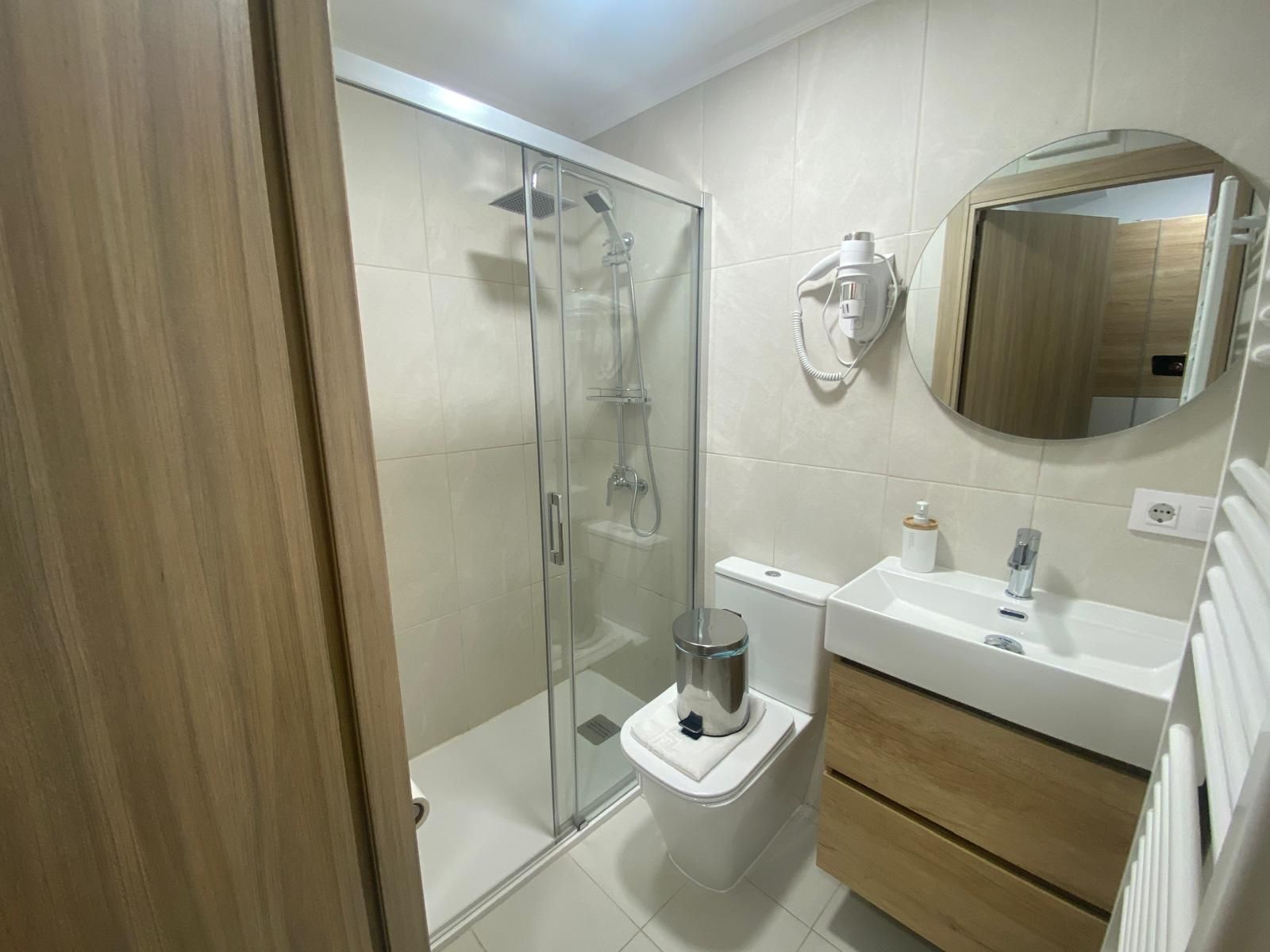 Baño moderno con ducha de cristal, inodoro, lavabo, tocador de madera y espejo de pared.